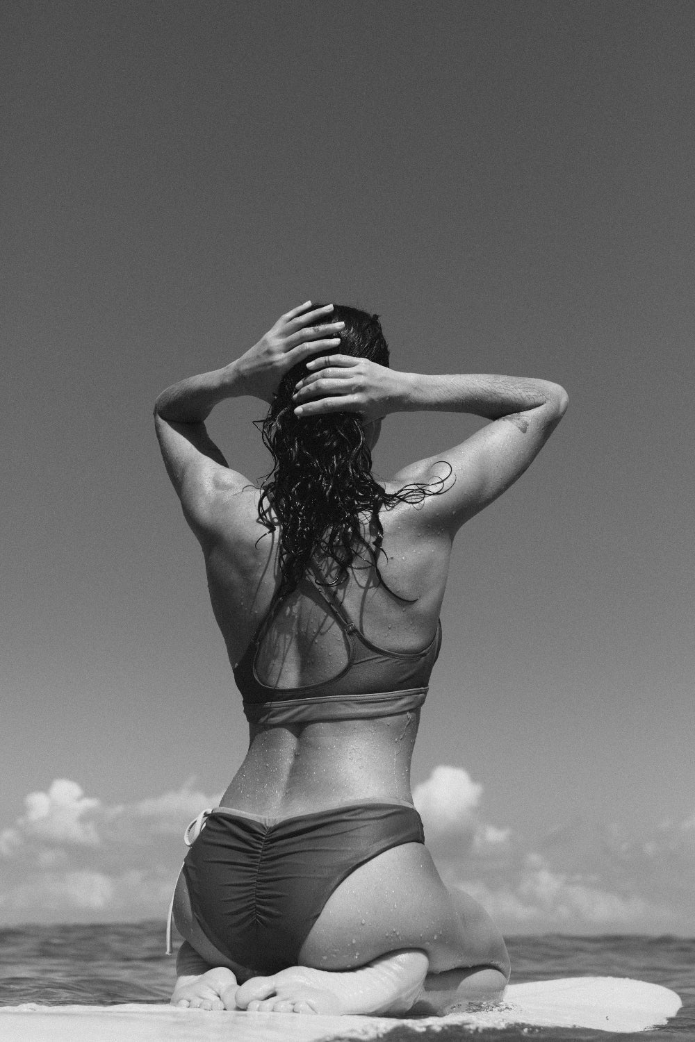 Woman in eco-friendly reversible bikini bottom kneeling on surfboard, hands in hair, with greyscale clouds and ocean behind