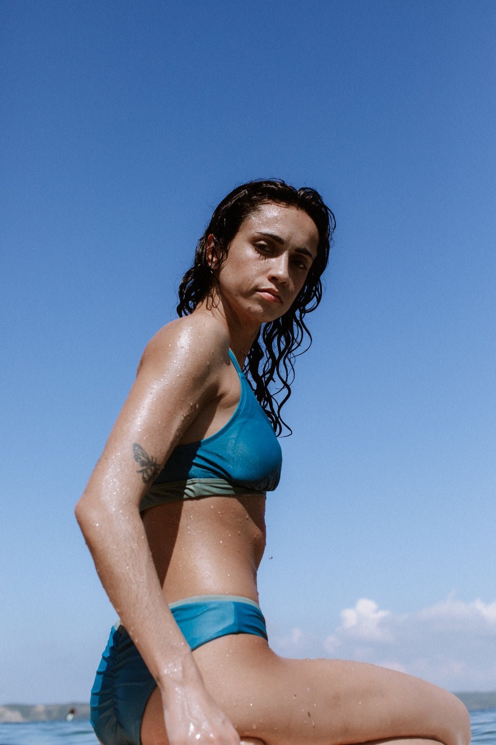 Eco-friendly drawstring bikini bottom styled for surfing, shown on wet skin under bright sun with blue ocean horizon