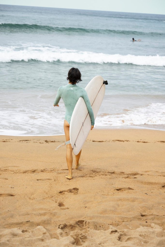 Surfer in aquamarine WALLIEN Yulex® wetsuit walking barefoot with surfboard on eco beach, facing ocean waves and sandy shore