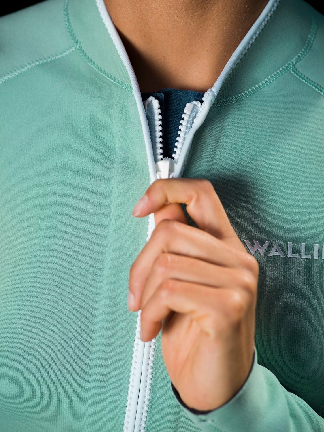 Close-up of woman zipping aquamarine Yulex® wetsuit with WALLIEN logo, showing eco-friendly texture and GBS stitching