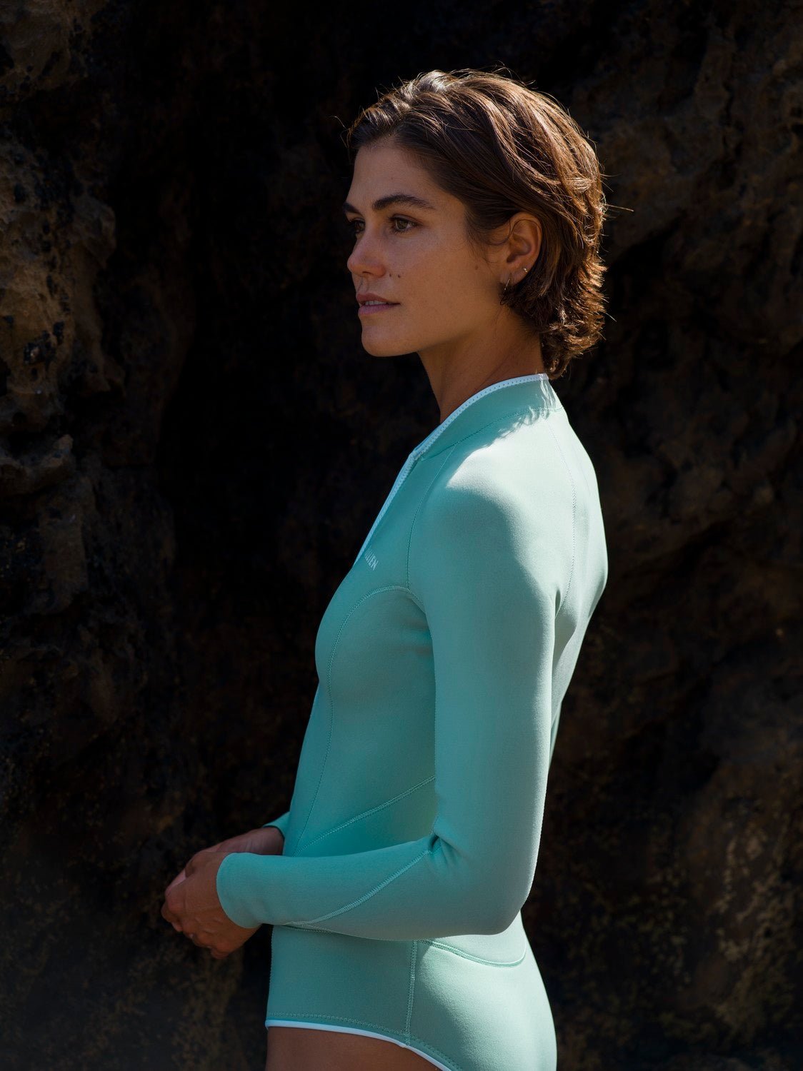 Side view of woman in eco-friendly WALLIEN Yulex® springsuit, standing near cliff face with natural rock textures behind