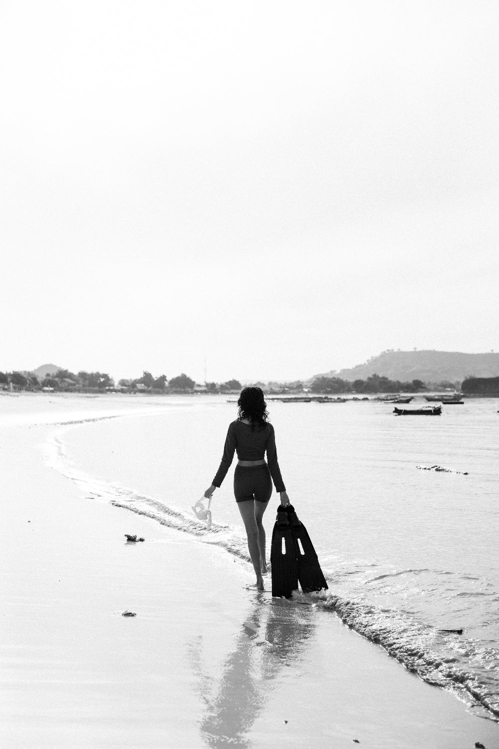 Woman in teal WALLIEN rash guard top walking into shallow surf with fins and mask, black and white shoreline in background