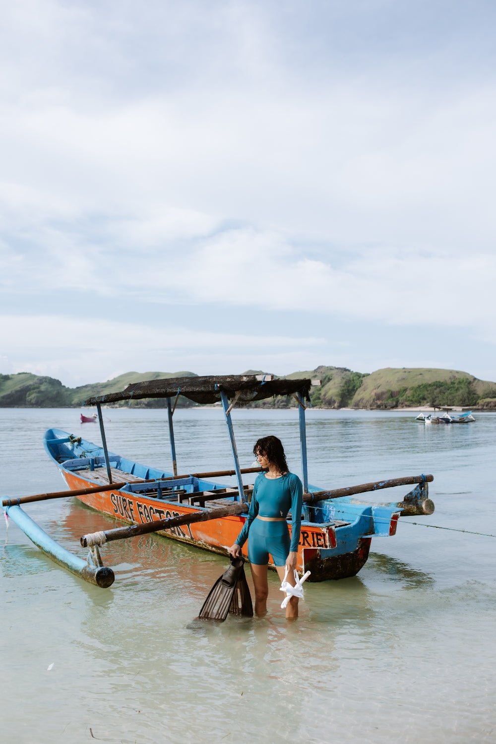 Woman in eco-friendly teal rash guard holding fins, stepping into ocean beside traditional fishing boat, island hills behind